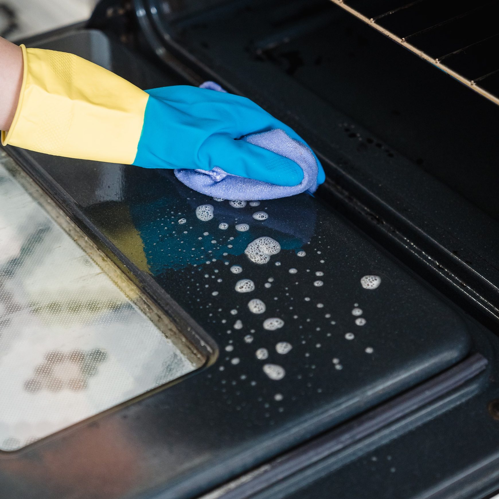 Blue-gloved hand wiping suds from an open oven door with a lilac cloth.