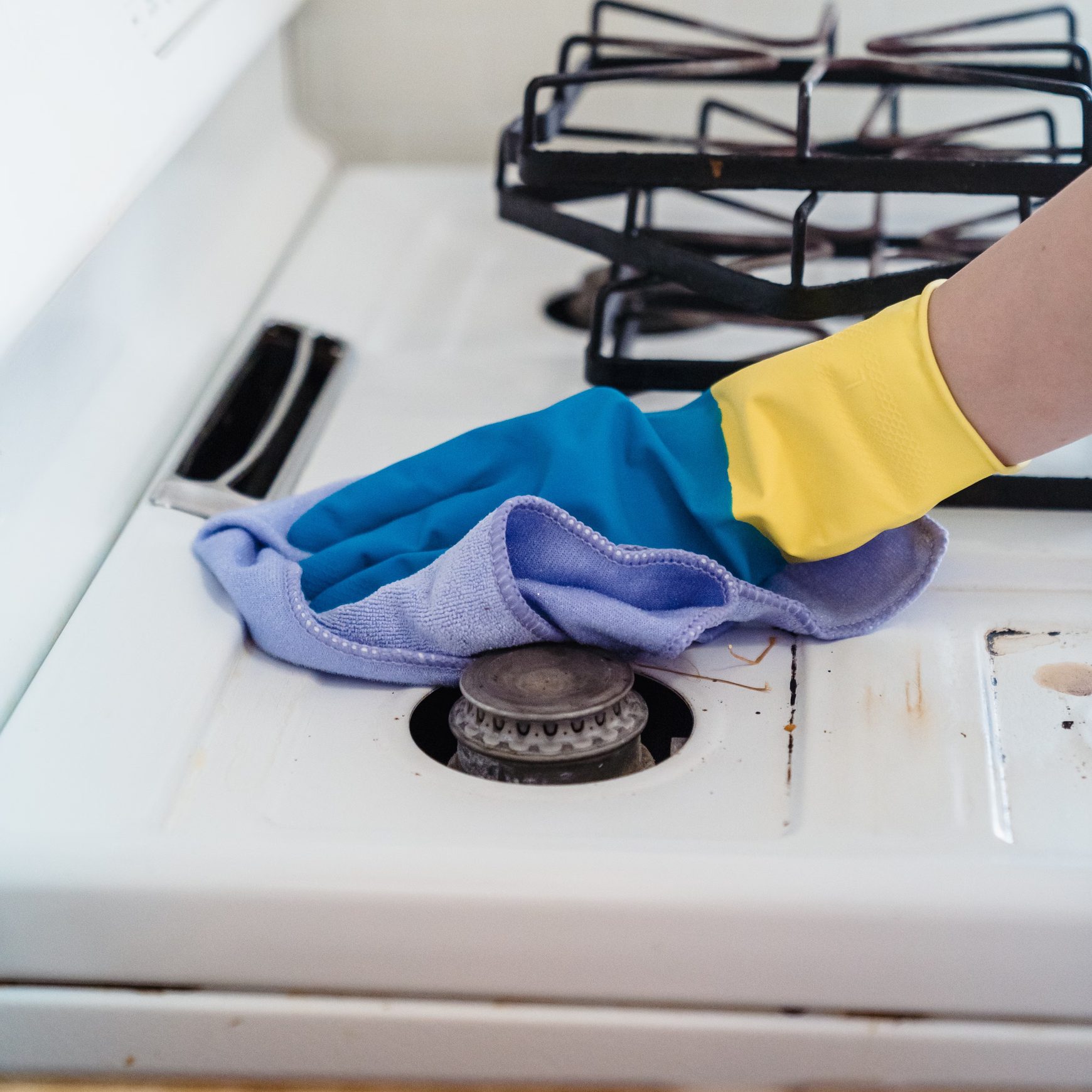 Blue-gloved hand wiping a white gas hob around a burner with a lilac cloth.