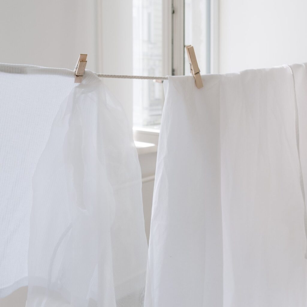 White fabrics hanging from wooden pegs on an indoor clothesline beside a bright window.