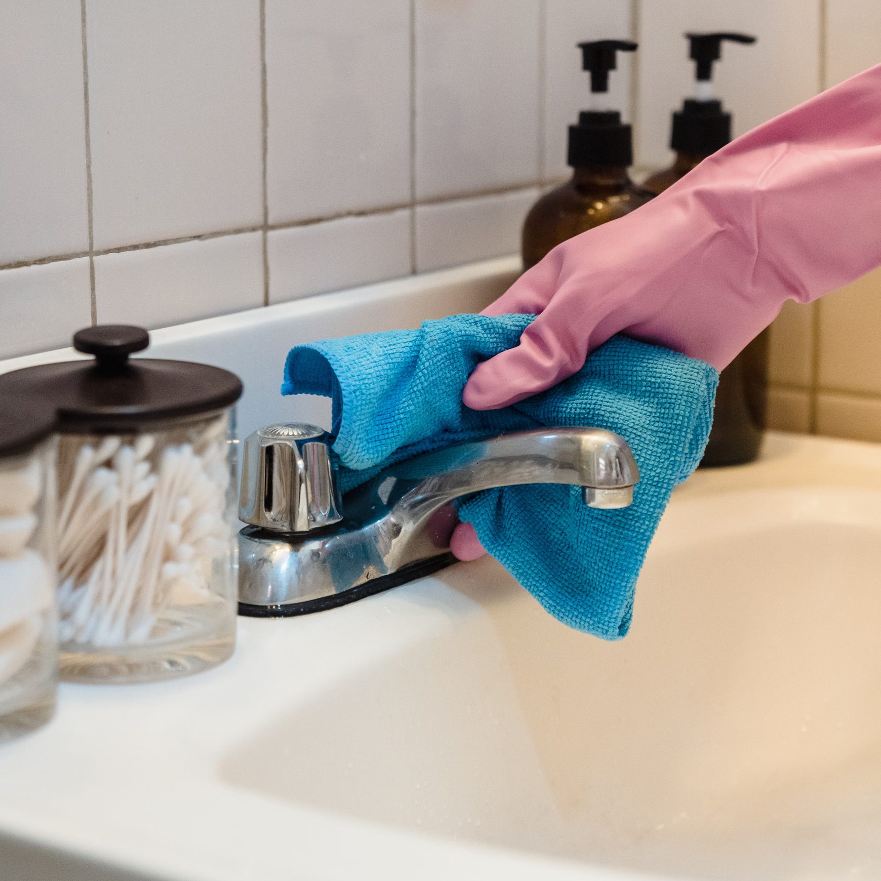 A pink-gloved hand wiping a bathroom tap with a blue cloth.