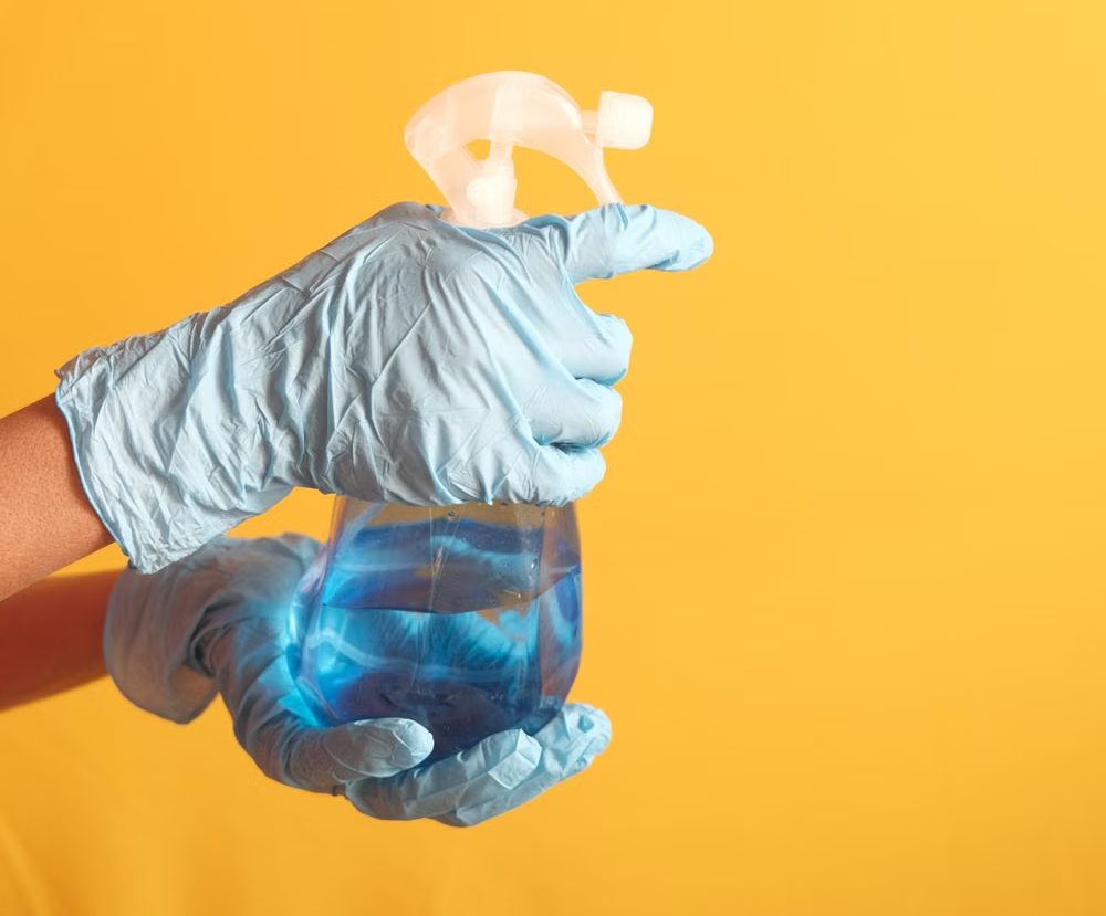 Blue-gloved hands holding a plain spray bottle against a yellow background.