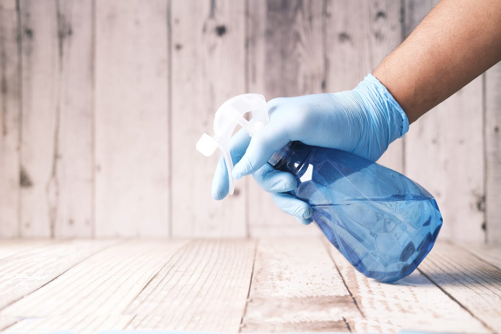 Blue-gloved hand holding an unlabelled spray bottle over a wooden surface