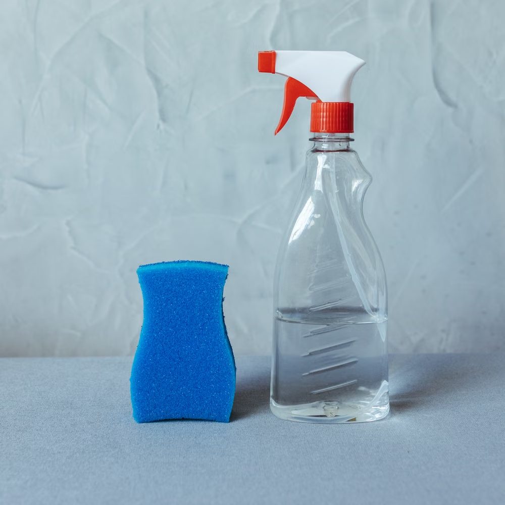 Plain spray bottle beside a blue cleaning sponge on a grey surface.