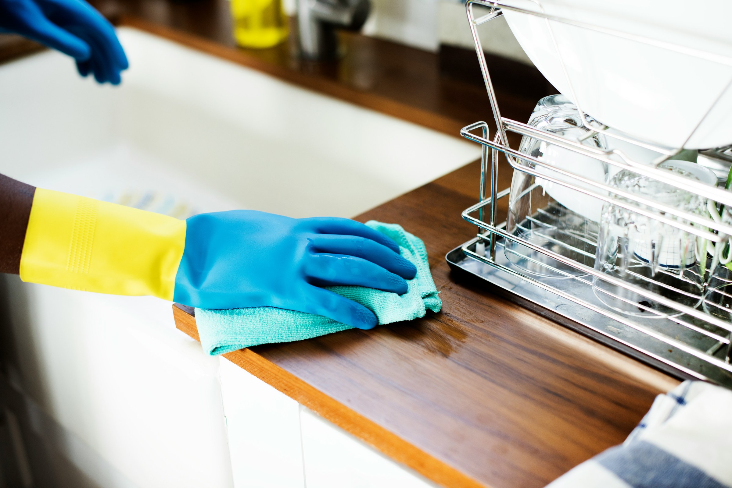Gloved hand wiping a kitchen worktop beside a dish rack.